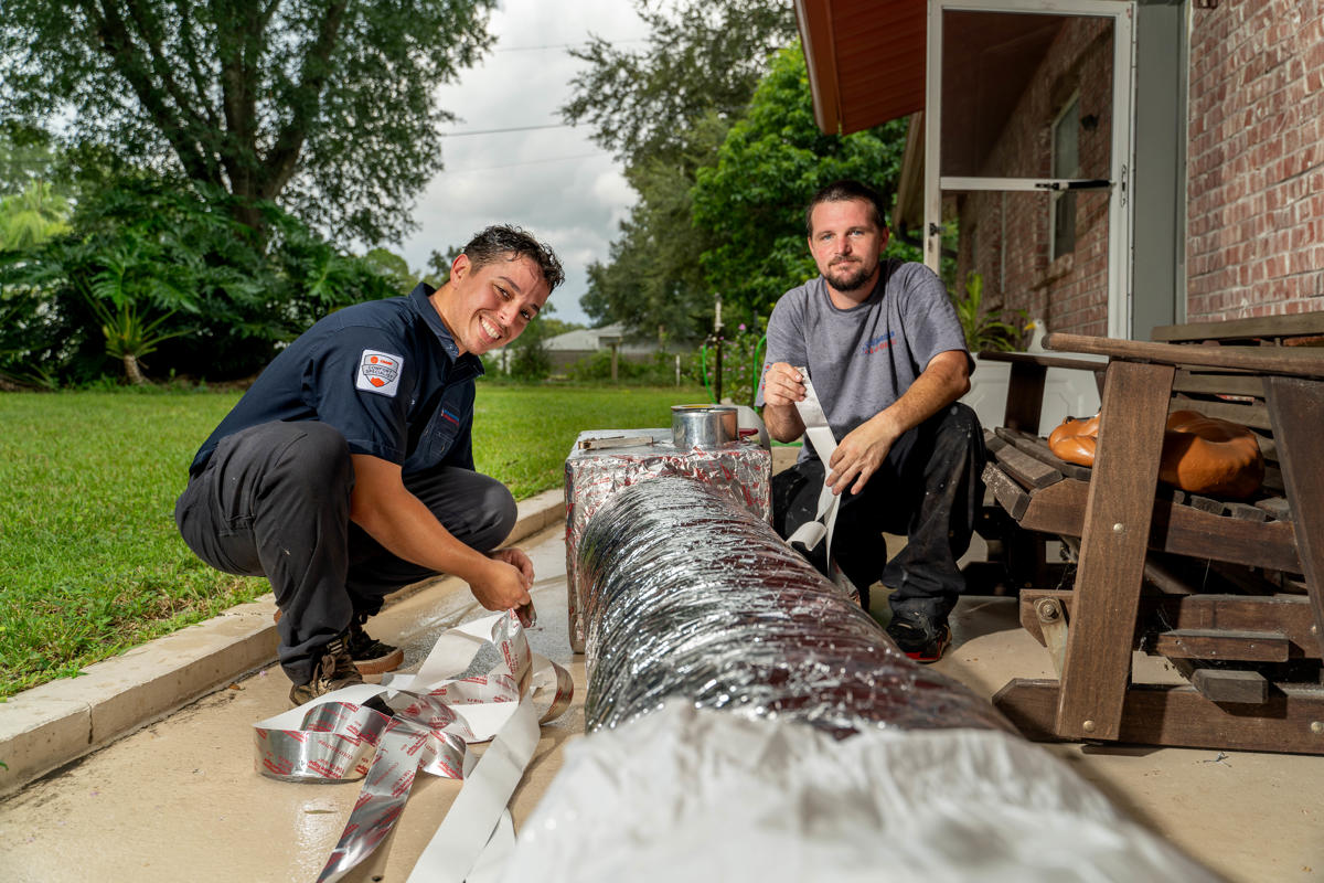 Air Conditioner Installation 5 Two Custom Air Conditioning Air Quality technicians work on wrapping and sealing an insulated air duct outside a home both smiling and focused on the task 1