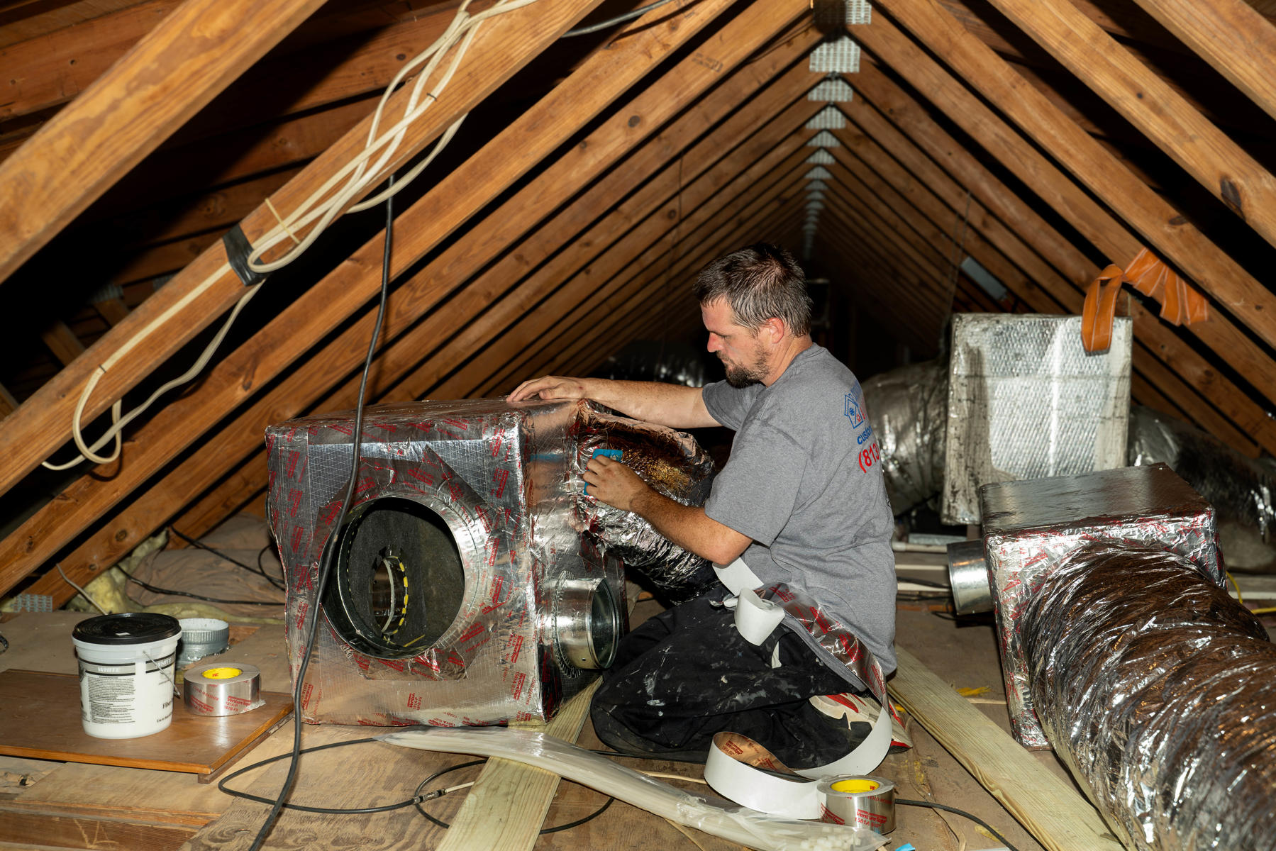 Ductwork 2 A technician works in a large attic sealing and insulating HVAC ductwork with metallic tape The technician is kneeling next to a large insulated air handler
